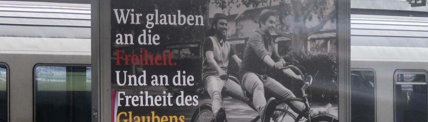 Plakat mit Muslim und Juden auf einem Tandem und Aufschrift: Wir glauben an die Freiheit. Und an die Freiheit des Glaubens. (Bundesjustizministerium)